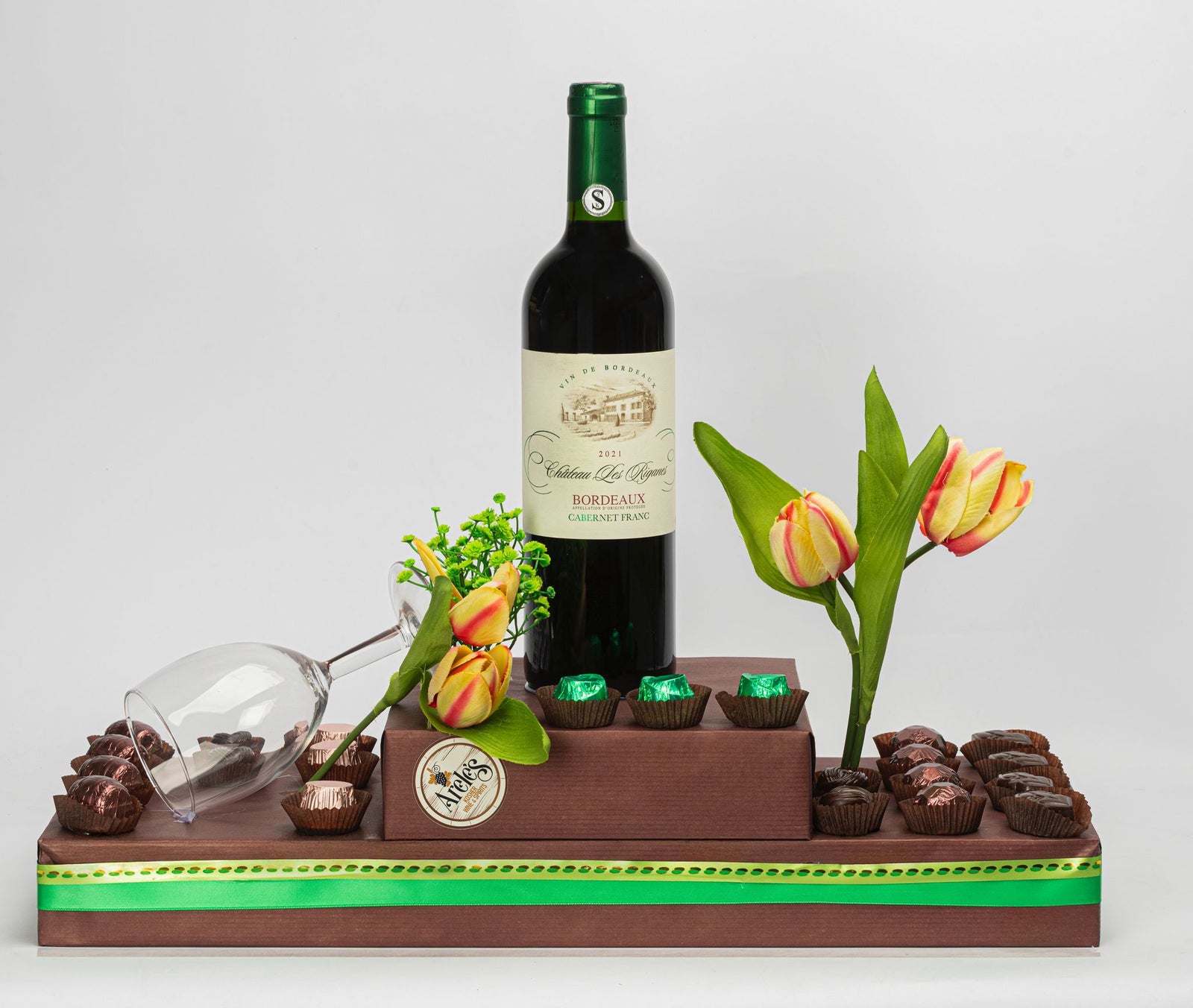 Image contains a kosher gift arrangement with glass wine cup, chocolates, tulips and a bottle of Chateau des Riganes - a dry red kosher wine. 